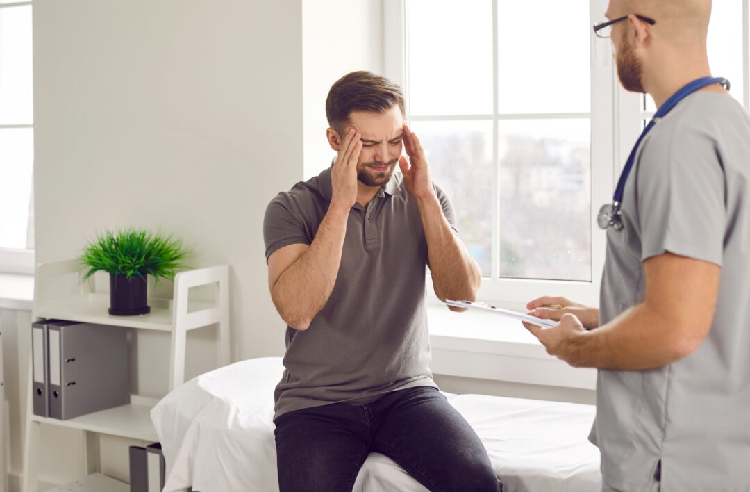 A man sitting in a doctor's office, holding his head in pain while a physician reviews paperwork, possibly discussing symptoms related to a brain injury. Contact a Missouri City brain injury lawyer for legal support after an accident.